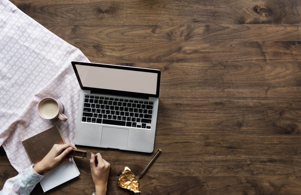 A person is using a laptop at a wooden table, with a coffee, a notebook, and a partially unwrapped chocolate bar. A patterned cloth is spread next to this person is getting ready to write a blog