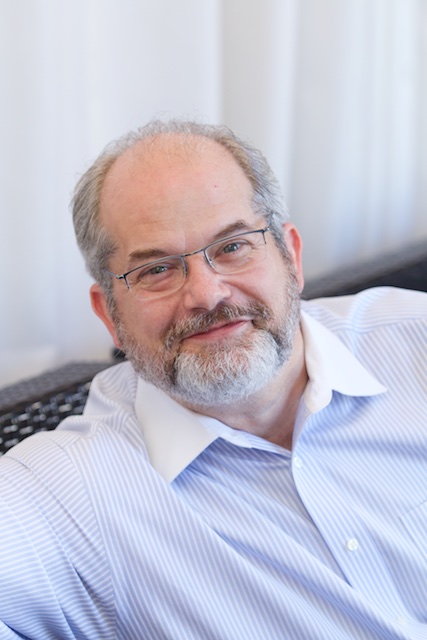 A man with a gray beard and glasses wearing a light blue shirt with white collar looks at the camera with a faint smile, sitting against a neutral background.