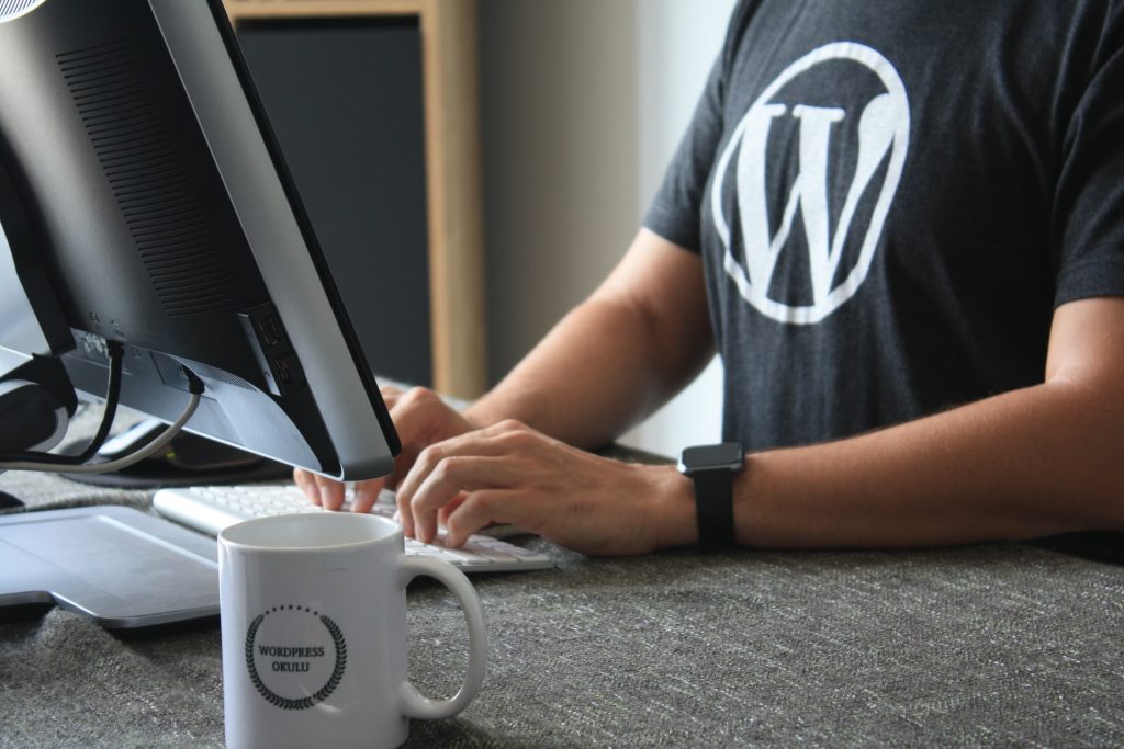 Person typing on a keyboard in front of a monitor, wearing a shirt with a WordPress logo. A mug with "WordPress.org" on it is on the desk.