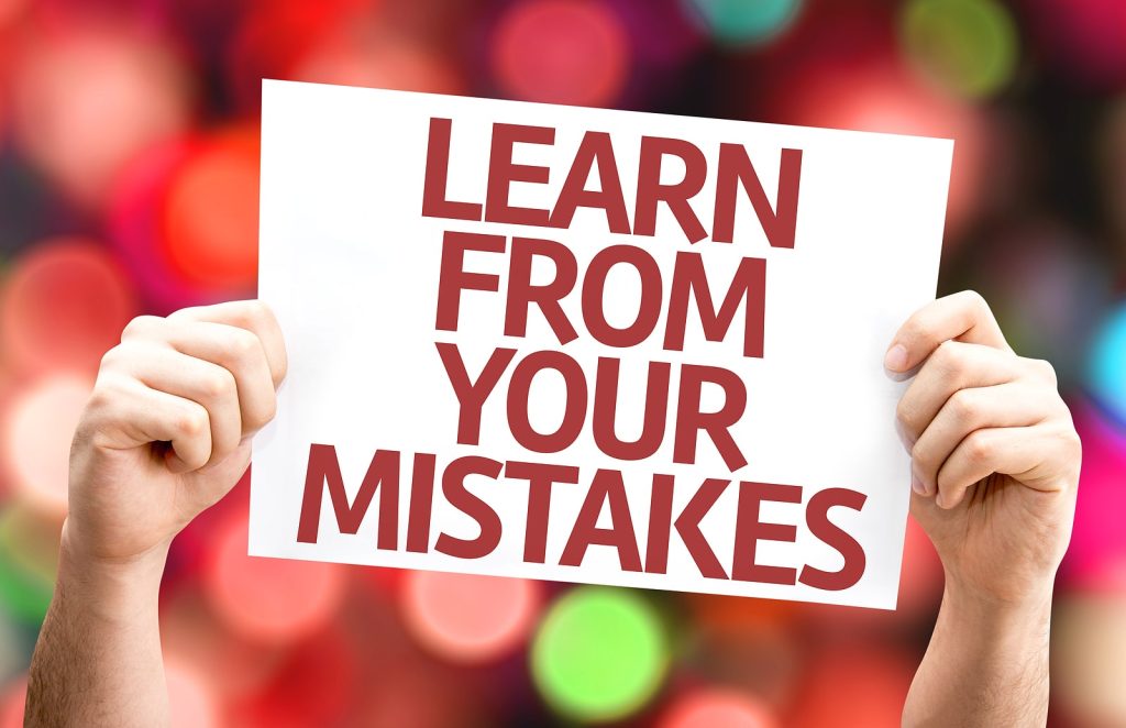 A person holding a sign that reads, "Learn from your mistakes," against a background of colorful, blurred lights, offering advice akin to blogging mistakes to avoid: lessons from experienced bloggers. -DM
