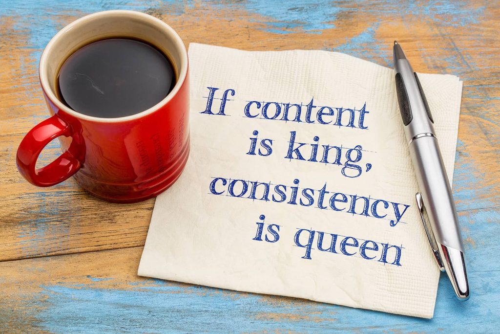 Red coffee cup, pen, and napkin with the text "If content is king, consistency is queen" on a painted wooden table.